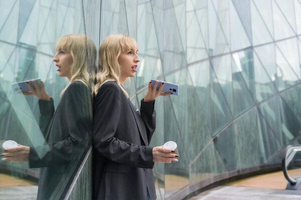 Entrepreneurial girl business park in a modern building reflected in the windows, young blonde in a suit sending a voice message with a take away coffee