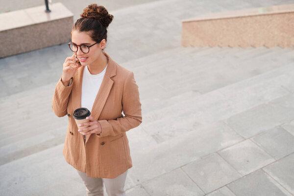 Above view of account manager in jacket drinking coffee and talking to client by phone being on move