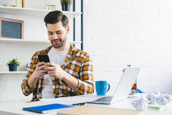 年轻的自由职业者在家里的办公室，坐在桌子旁使用智能手机工作。