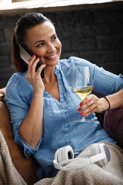 Leisure time concept. Happy beautiful woman talks on a phone and drinks white wine from glass sitting on a couch indoors. Female spending her free day and relaxing at home alone