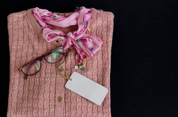 Stylish pink women's sweater on black surface with blank label,reading glasses and authentic scarf.Special days concept.