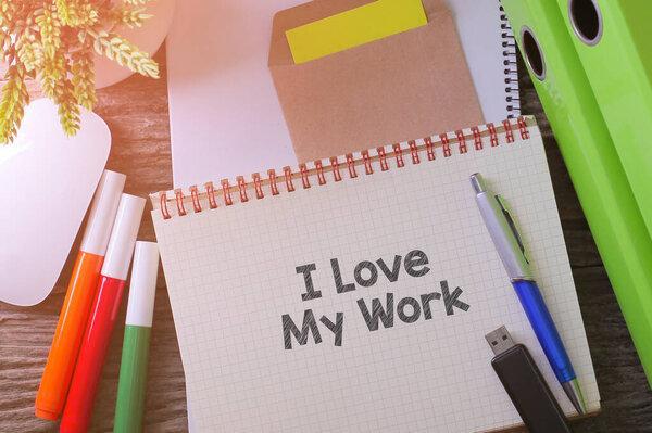 Notebook writing "I Love My Work" on table with Working space at the office with plant, usb drive and glasses