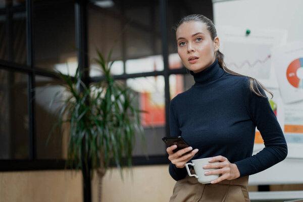 young confident brown-haired woman holding a phone and a cup of 