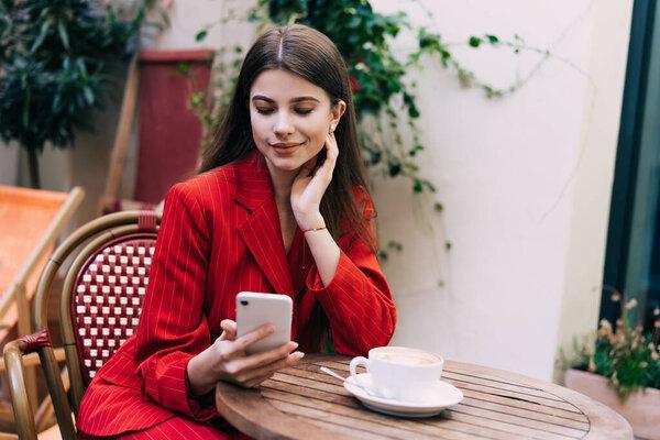 Happy young female in stylish outfit messaging on cellphone while smiling and sitting at table with hand on chin pose at cozy cafe