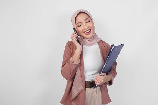 Young Asian Muslim woman wearing hijab talking on a phone call while holding document over isolated white background.