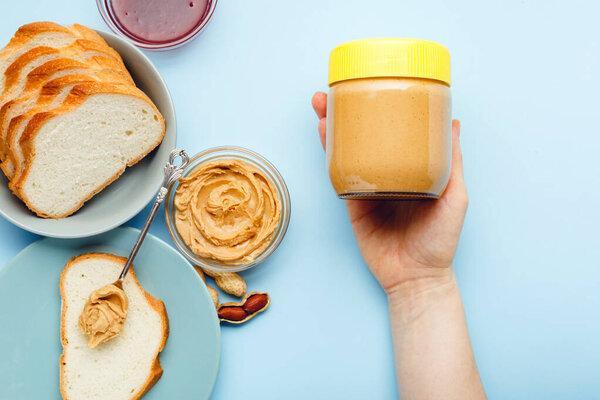 Creamy peanut paste in glass jar with yellow cap in female hand and peanuts in the peel scattered on blue colored background. Cooking breakfast with peanut butter. Vegan food concept.