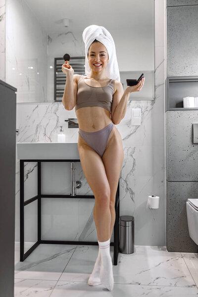 全长的年轻女性化妆刷看着相机，同时在家里的浴室里摆姿势。美容美发概念
