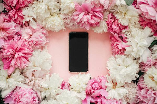 Pink and white peonies and phone with blank screen on pastel pink paper. Stylish flat lay with copy space. Happy mothers day, floral greeting card mockup. International Womens Day.