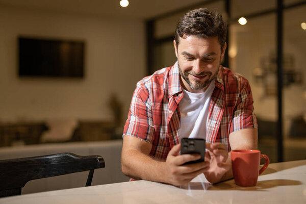 Handsome man with mobile phone sitting on weekend at home. American man using phone while sitting on sofa at living room interior. Relaxing text with smartphone. Call phone