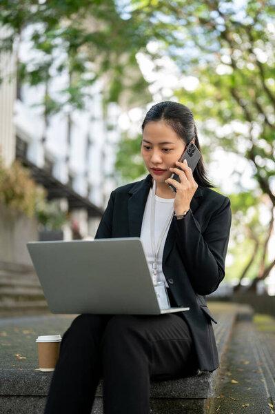 A busy and professional millennial Asian businesswoman is having a business phone call while working on her laptop on the stairs in the city. Urban busy lifestyle and corporate job concept