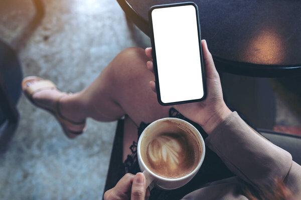 Top view mockup image of woman's hands holding black mobile phone with blank white screen while sitting and drinking coffee in cafe