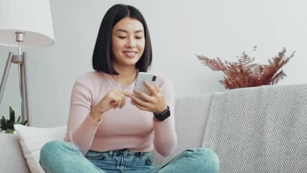 年轻的亚洲女人在手机上浏览社交媒体，享受着惊人的新闻，坐在家里的沙发上