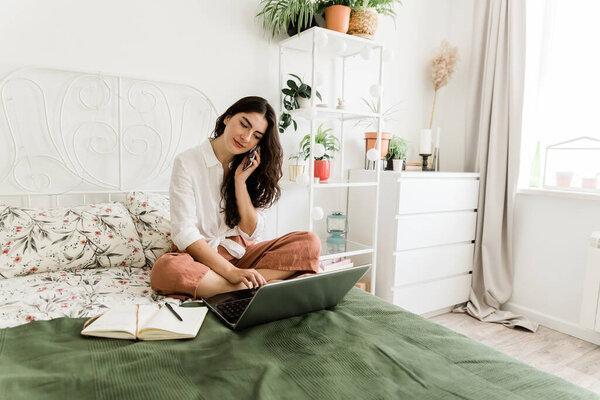 Woman sitting on bed talking on the phone and working using laptop.