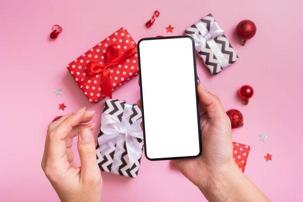 Online Christmas shopping concept. Female hand with phone mock up and Christmas presents flat lay on a pink background.