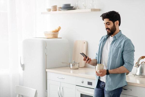 Morning routine. Young arab man using smartphone and drinking coffee in kitchen, free space