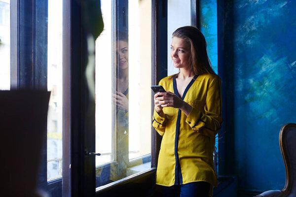 Pensive girl using mobile telephone while leaning on the window 
