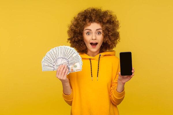 Wow, online banking, money transfer! Portrait of surprised curly-haired woman in urban style hoodie showing dollar banknotes and cellphone, looking with amazement. indoor studio shot, isolated