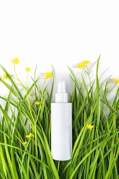 One white spray bottle with cosmetic product among the green grass, yellow flowers on white background. Natural Organic Spa Cosmetic concept Mockup.