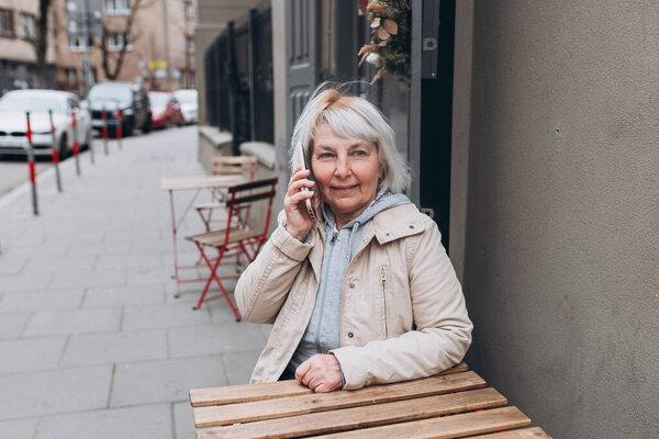 Mature woman is sitting in cafe and relaxing. Senior is talking on phone outdoors. Attractive 60s female having conversation. Urban lifestyle concept
