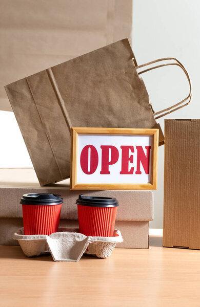A lot of Craft Paper Bags and Boxes stay near Cups of Coffee with sign Open. Concept of Delivery Service Packing Order Take Away for Customer.