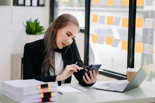 Young beautiful woman talking on smartphone, using calculator and laptop computer while sitting at working place. Concentrated at work in modern office