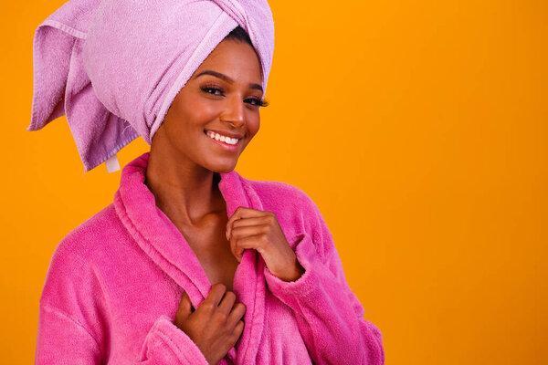 Afro woman with towel on her head and bathrobe after getting out of the bath on yellow background. Bath and care concept