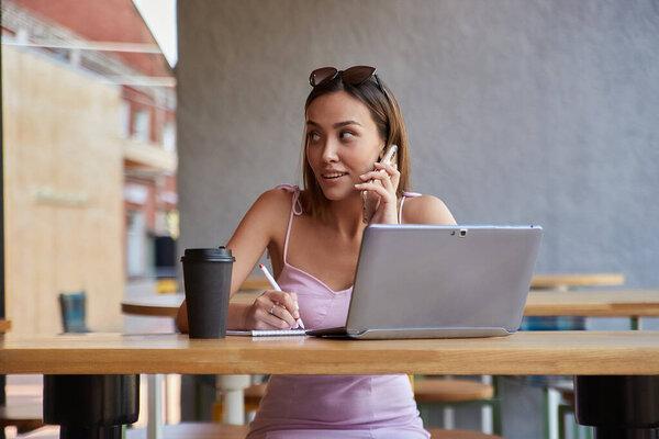 beautiful young asian woman sitting with laptop at cafe, calling by smartphone