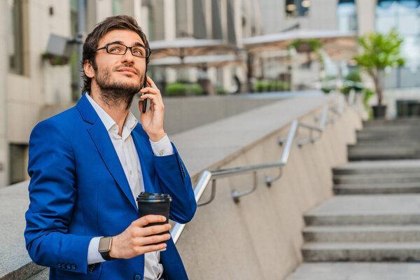 Attractive young businessman in suit standing outdoors at the city street with takeaway coffee cup while talking on mobile phone with look away