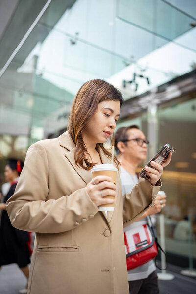 年轻的亚洲优雅的商界女性领导者身穿西服，站在现代城市建筑中，使用手机平台应用程序，在街上边走边喝着咖啡.