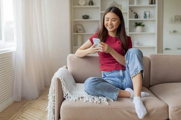 Great App. Smiling Young Asian Woman Browsing Application On Smartphone While Sitting On Couch At Home, Happy Korean Female Using Mobile Phone While Relaxing In Cozy Living Room Interior, Copy Space