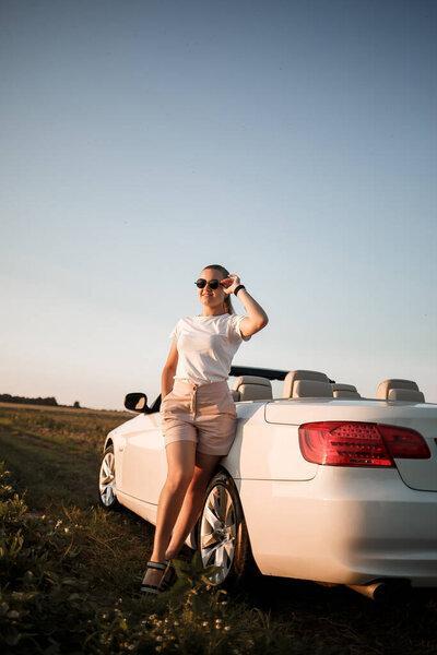 A beautiful young woman of European appearance on the face of sunglasses, she stands near her white convertible. Successful woman businesswoman and her white car.