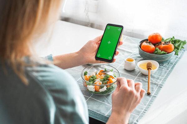Concept of calorie counting, dietary nutrition online shopping and online education. A blonde woman has breakfast with a healthy vitamin salad and holds a phone with a green chroma key screen.