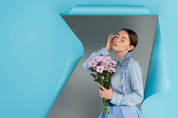 elegant woman touching face while posing with closed eyes and pink flowers near blue torn paper on grey background