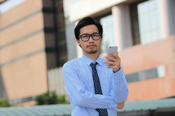 Portrait of handsome young Asian business man holding mobile smart phone and looking on camera at outside office background.