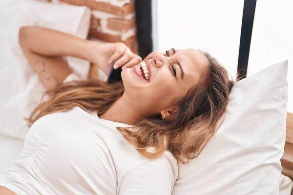 年轻美丽的惊慌失措的女人躺在床上用智能手机说话