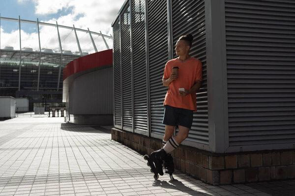 Joyful roller skater holding smartphone and coffee to go near building on urban street 