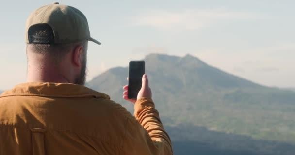 一个留着胡子的男人带着博客旅行，一边在巴厘岛山区旅行，一边用手机拍摄火山的景象。一位博主记录了他的冒险经历.