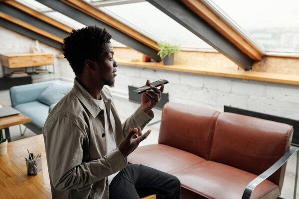 Side view of young African American male employee recording voice message in office while holding smartphone in front of his face