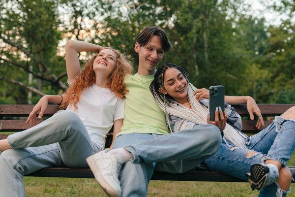 Three happy young teenagers friends having fun at the park, sitting on a bench taking a self portrait with a cellphone. School education. Youth lifestyle.