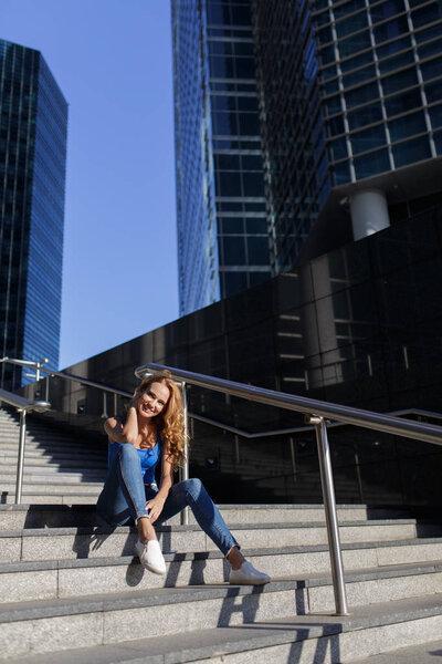 Beautiful woman sitting on the steps between skyscrapers with her smartphone