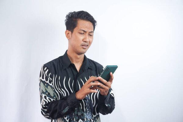 当看到智能手机时，年轻的亚洲男人哭了，伤心了。身穿蜡染衬衫的印度尼西亚男子.