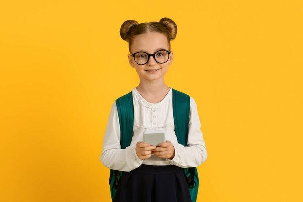 带着智能手机、背包带黄色工作室背景的可爱小女孩的肖像、使用手机、在相机前微笑、复制空间