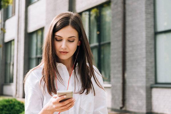 attractive young woman in white shirt holding smartphone and listening music in earphones on street