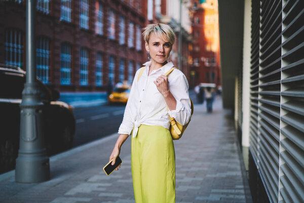 Half length portrait of beautiful female student dressed in yellow apparel looking at camera while standing on street of modern urban setting, young woman with stylish short haircut posing outdoors