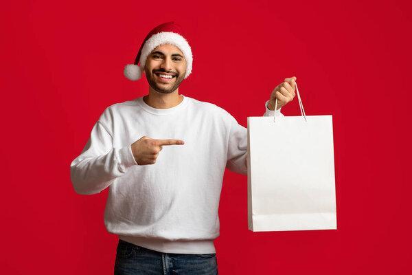 Cheerful Arab Guy Wearing Santa Hat Pointing At Blank Paper Shopping Bag
