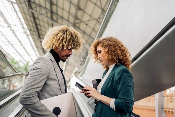 Business woman with formal wear using her mobile phone and talking with her african coworker in train station.