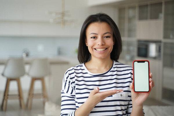 Happy young woman holding phone with copy space, advertising new mobile apps. Smiling modern female showing smartphone with mockup empty screen, looking at camera, standing at home. Online store offer
