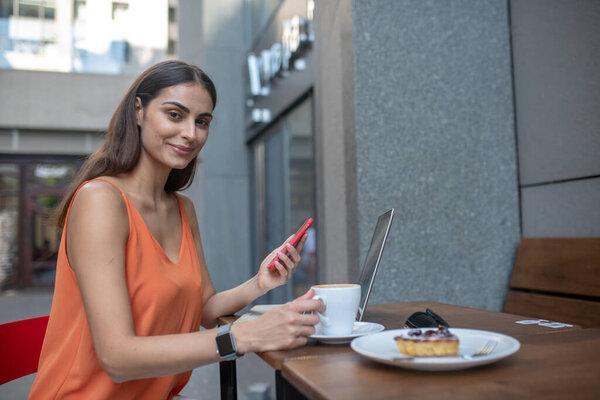 Trendy cute young woman holding smartphone and having coffee
