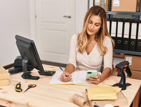 Young blonde woman ecommerce business worker using smartphone and writing on book at office