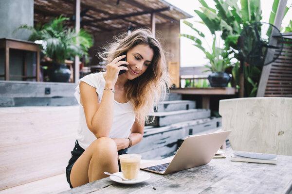 Beautiful young woman in white t-shirt and black shorts sitting at table, speaking by mobile phone and watching laptop on summer day